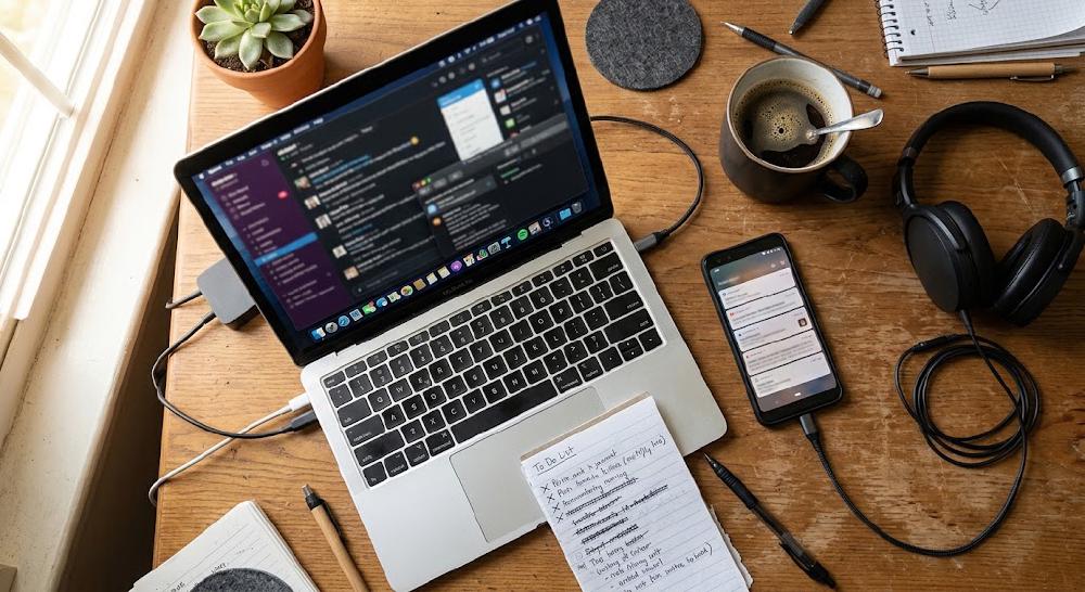 Desk from above: laptop with many open chat and mail windows, a smartphone full of notifications beside it, headphones, coffee and a handwritten to-do list